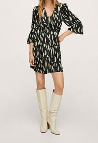 Schwarzes Kleid mit abstraktem cremfarbenen Muster, V-Ausschnitt und ausgestellten Ärmeln. Kombiniert mit knielangen weißen Stiefeln und einer glatten Textur.