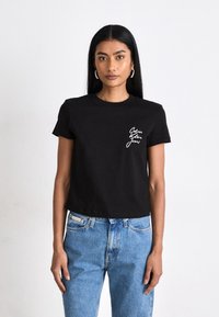 Camiseta de algodón negra de manga corta con cuello redondo, con texto de logo blanco en el lado izquierdo del pecho, combinada con jeans de mezclilla azul claro.