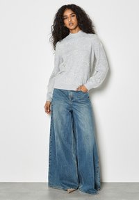Hellgrauer, gestrickter Pullover mit Schleifenakzenten, kombiniert mit weiten, blauen Jeanshosen. Lässige Passform mit gerippten Bündchen und Saum.