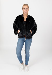 Giacca bomber in pelliccia sintetica nera con chiusura a zip, polsini e vita a coste. Indossata sopra un top nero, abbinata a jeans skinny blu e sneakers bianche.