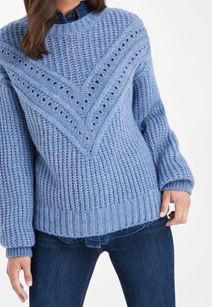 Femme portant un pull bleu en maille épaisse avec un motif ajouré en chevron, superposé à une chemise en denim et associé à un jean foncé.