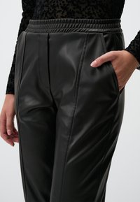 Schwarze Kunstlederhosen mit elastischem Bund, vorderen Falten und Seitentaschen. Der Stoff hat eine glatte, glänzende Textur.