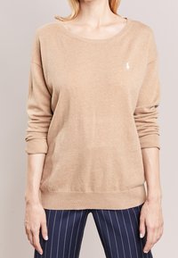 Beżowy sweter dziany z szerokim dekoltem i ściągaczami. Posiada małe białe logo na lewej piersi. Miękka tekstura i luźny krój.