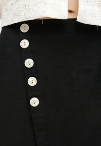 Prim-plan al unei fuste negre cu cinci nasturi albi pe lateral, purtată cu un top alb din dantelă care lasă la vedere o parte a taliei.