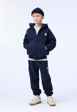 Granatowa hoodie na zamek błyskawiczny z logo obcego, w zestawie z dopasowanymi dresami. Tkanina wydaje się miękka; ma mankiety i ściągacz w pasie. Pod spodem biała koszulka.