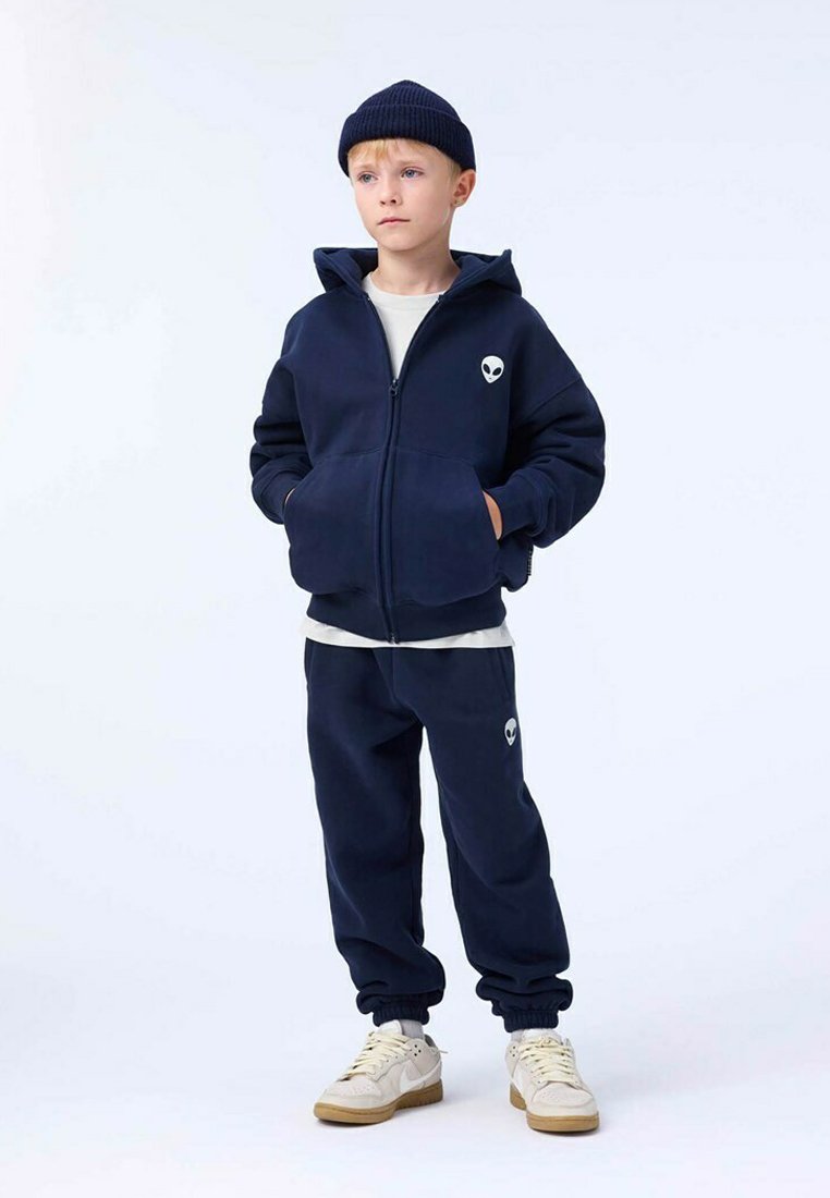 Námořnická modrá zipová mikina s logem mimozemšťana, ve spojení s odpovídajícími tepláky. Tkanina vypadá jemně; má manžety na rukávech a pasu. Bílá tričko pod tím.