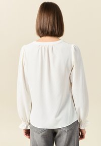 Camicia bianca con maniche a sbuffo lunghe, scollo arricciato e polsini elasticizzati. Tessuto liscio, vestibilità comoda, abbinata a jeans grigi.