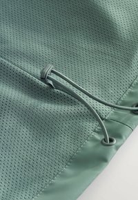 Țesătură verde deschis cu textură de plasă, având un sistem de închidere cu sfoară și un toggle gri, precum și ochiuri pe margine.