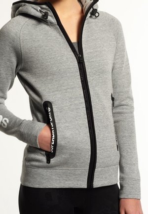 Sweat à capuche gris zippé avec bordure noire, capuche et poches zippées ; une main dans la poche et texte blanc du logo sur la manche gauche et le bord de la poche.