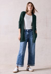 Lång grön cardigan i mjukt stickat material, lagd över en vit topp, tillsammans med vida blå denimjeans och vita sneakers med svarta detaljer.