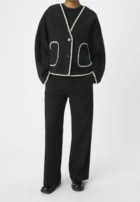 Person i svarta vida byxor och en svart knappad kofta med vit zickzackkant på fickor och kanter.