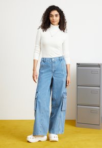 Lysvasket denim cargo bukser med brede ben og lommer, parret med en hvid ribbet turtleneck, sat mod en gul baggrund.