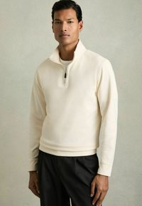 Hellcremefarbener Half-Zip-Pullover aus weichem Stoff, mit gerippter Textur, langen Ärmeln und einem schlichten schwarzen Reißverschlussdetail.