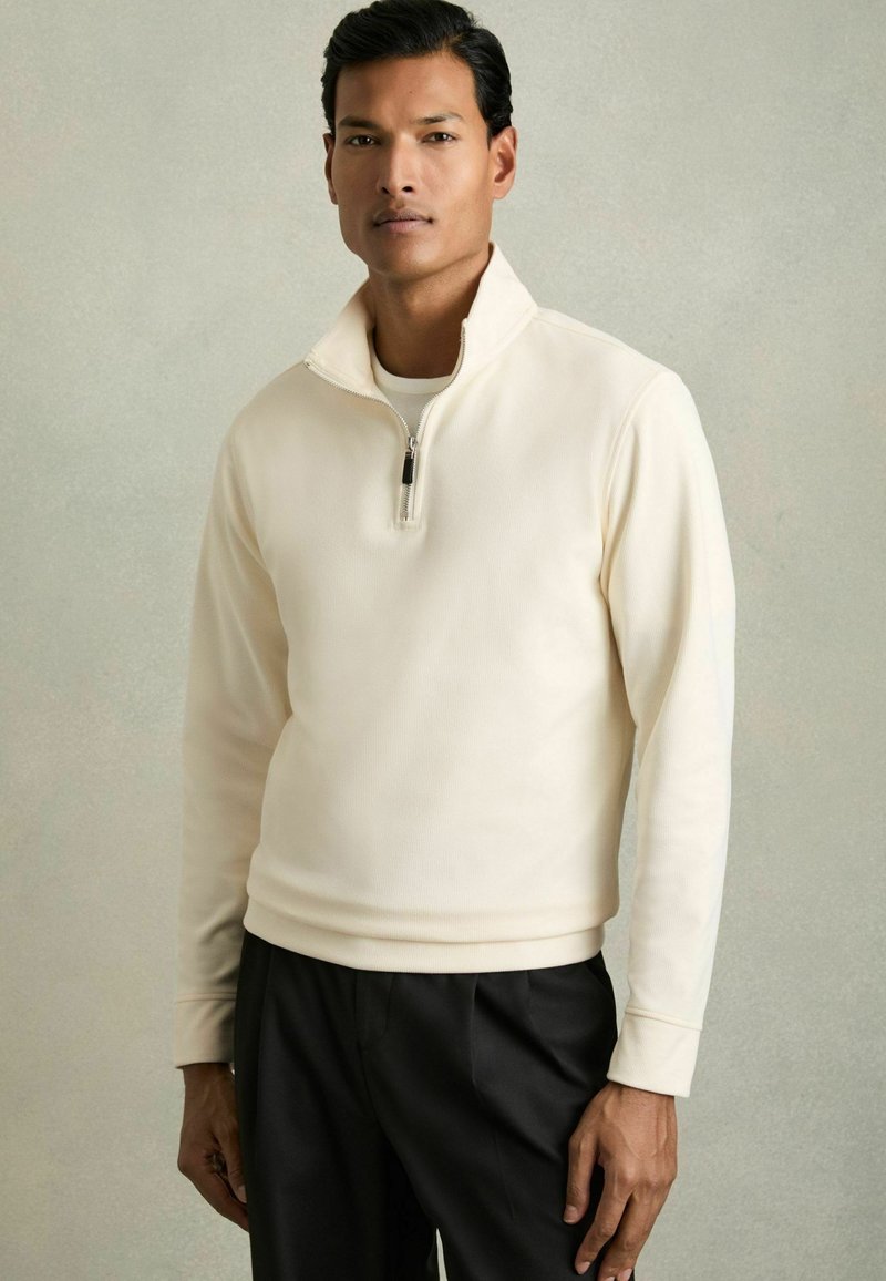 Hellcremefarbener Half-Zip-Pullover aus weichem Stoff, mit gerippter Textur, langen Ärmeln und einem schlichten schwarzen Reißverschlussdetail.