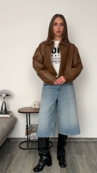 Ruskea nahkatakki, valkoinen graafinen t-paita, vaaleansiniset väljät farkkushortsit ja mustat polven yli ulottuvat saappaat neutraalissa ympäristössä.