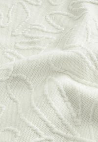 Tissu blanc texturé avec motifs floraux et feuilles en relief cousus, montrant des plis doux et une surface tissée détaillée.