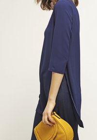 Mörkblå, långärmad topp med lös passform, som har en rundad nederkant och en slits baktill. Kommer med en gul, flätad handväska.