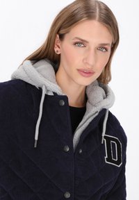 Granatowa sztruksowa kurtka z pikowanym wzorem, szarym polarem wewnątrz, kapturem ze sznurkiem i czarną naszywką z literą "D" na piersi. Srebrne elementy wykończeniowe.