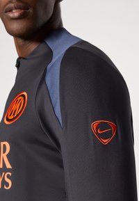 Crna atletska majica s narančastim timskim logotipom i narančastim Nike znakom na rukavu. Sadrži plave naglaske na ramenu.