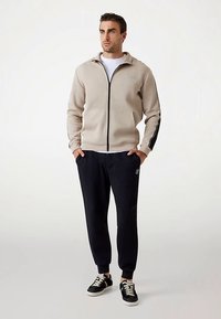 Beige jacka med dragkedja och svarta detaljer, vit t-shirt och svarta joggingbyxor. Sneakers med svarta och krämfärgade designelement.