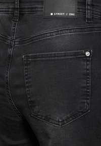 Zwarte denim jeans met een achterzak voorzien van een zilveren knoopdetail. De stof heeft een soepele textuur en subtiele vervaging.