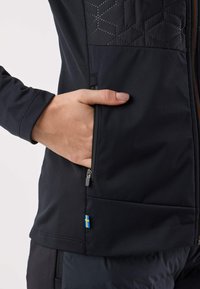 Giacca softshell nera con motivo trapuntato esagonale sulla parte superiore del petto, tasca laterale con zip e un piccolo dettaglio con la bandiera svedese sull'orlo.