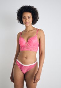 Rosa spetslingerie-set med en bralette med vågiga kanter och blommönster, i kombination med matchande stringtrosor och ett vitt midjeband.