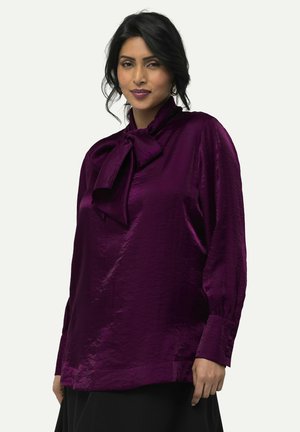 Blouse en satin violet avec un col haut et un grand nœud décoratif, manches longues avec des poignets boutonnés et une finition lisse.
