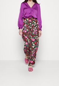 Blusa di raso viola con scollo a V e bottoni, abbinata a pantaloni neri fluidi coperti da fantasie floreali colorate e sandali rosa con punta aperta.
