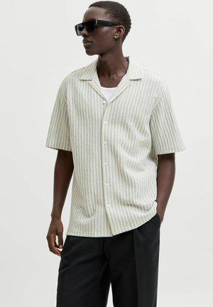 Homme portant une chemise texturée à manches courtes boutonnée, un pantalon noir à jambes larges, des lunettes de soleil foncées, et debout avec une main dans la poche.