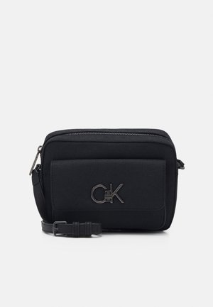 Zwarte compacte crossbodytas met voorvak, zilverkleurig Calvin Klein-logo, ritssluiting en verstelbare band op een effen achtergrond.