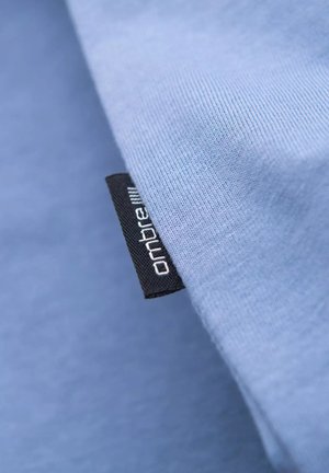 Light blue fabric with a smooth texture. Features a black tag with "ombre" printed in white, partially visible at the edge.