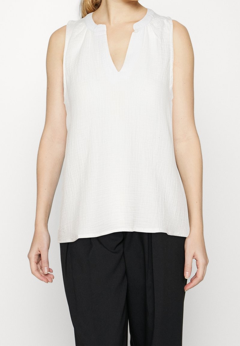 Femme portant un chemisier blanc texturé sans manches avec un col en V en encoche et un pantalon noir ample, debout devant un fond uni.