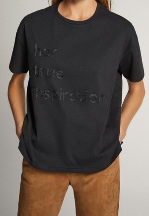 Black short-sleeve T-shirt with the embroidered text "her true inspiration," worn by a person with hands in brown pants pockets.