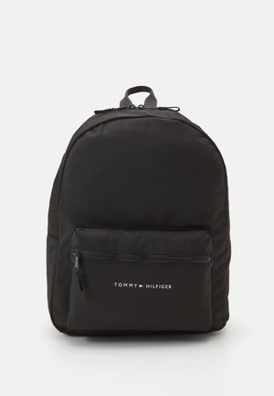 Sac à dos noir avec une forme arrondie, fabriqué en tissu lisse. Il comporte une poche zippée à l'avant et un logo imprimé en blanc et rouge. Poignée sur le dessus incluse.