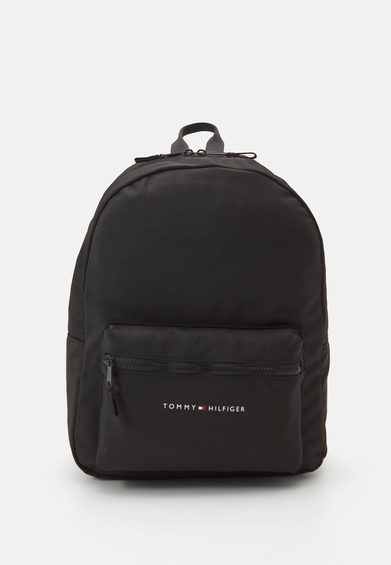 Mochila preta com formato arredondado, feita de tecido liso. Apresenta um bolso frontal com fecho e um logótipo impresso em branco e vermelho. Inclui uma pega na parte de cima.