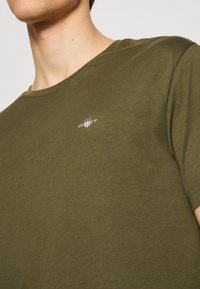 T-shirt verde oliva realizzato in morbido cotone, con scollo rotondo e un piccolo logo con accenti rossi, bianchi e blu sul petto.