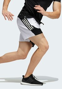 Musta ja valkoinen urheilushortsit, joissa on kolme pystysuoraa mustaa raitaa, yhdistettynä mustiin juoksukenkiin. Keskitä liikkeeseen ja muotoiluun.