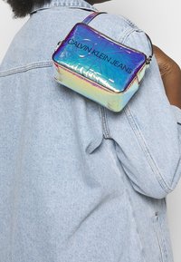 Irisfärgad rektangulär väska med dragkedja, med texten "CALVIN KLEIN JEANS" i svart och en tunn justerbar rem.