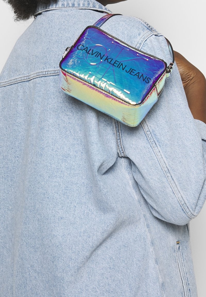 Borsa rettangolare iridescente con cerniera, con il marchio "CALVIN KLEIN JEANS" in testo nero e una sottile tracolla regolabile.