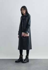 Femme debout portant un long manteau en cuir noir, des leggings noirs, des bottes montantes noires à lacets, et tenant un petit sac à main rose devant un fond uni.