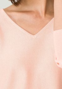 Gros plan sur une personne portant un pull en V rose pâle, montrant le décolleté et une partie du bras levé près du visage.