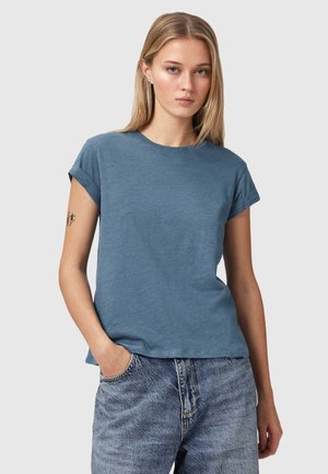 Ung kvinde med langt blondt hår iført en blå t-shirt med opsmøgede ærmer og lyseblå jeans i denim med den ene hånd i lommen.