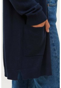 Navy gestrickte Strickjacke mit gerippten Bündchen und seitlichen Schlitzen, versehen mit einer einzelnen Tasche. Kombiniert mit leicht strukturiertem blauen Denim.
