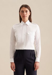 Camicia bianca a maniche lunghe con chiusura a bottoni, due tasche sul petto e colletto strutturato, abbinata a pantaloni scuri, tessuto di cotone liscio.