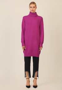 Übergroßer magentafarbener Strick-Turtleneck-Pullover, kombiniert mit schwarzen Jeans, die Fransen und metallische Akzente an den Bündchen aufweisen. Schwarze Absätze runden das Outfit ab.