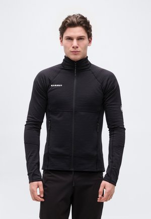 Fiatal férfi, aki testhez simuló fekete cipzáras dzsekit visel, melyen Mammut logó található a mellkason és a bal ujján, egyszínű világos háttér előtt áll.