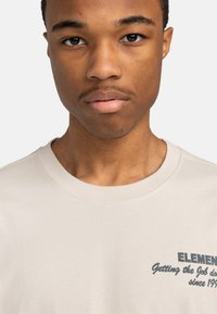 Béžové bavlněné tričko s kulatým výstřihem; má tmavě modrý text na hrudi, který říká "ELEMENT" a "Od roku 1992 plníme úkoly."