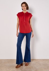 Rote Bluse mit kurzen Ärmeln, einem Reißverschluss vorne und zwei Brusttaschen, kombiniert mit blauen Schlagjeans und braunen Plateauschuhen.