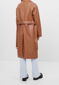 Hnědý kožený trenčkot s opaskem, dlouhými rukávy a středním rozparkem, spárovaný se světle modrými zvonovými džínami a silnými černými botami.